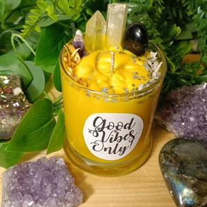 May include: A yellow candle with a clear glass jar and a white label that says "Good Vibes Only". The candle is surrounded by crystals and greenery.