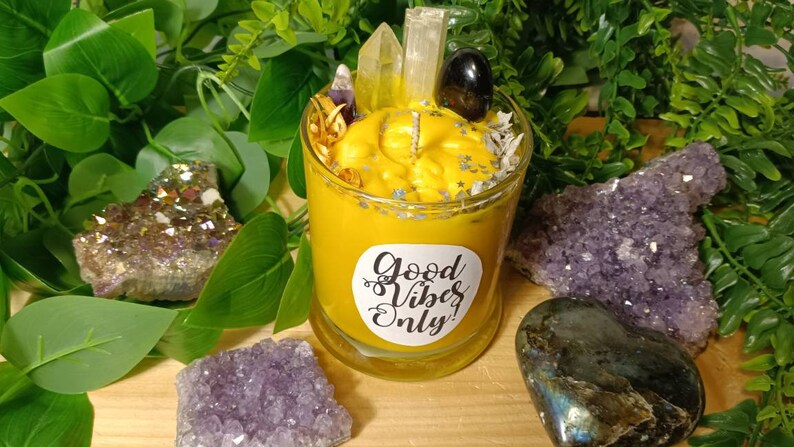 May include: A yellow candle with a clear glass jar and a white label that says "Good Vibes Only". The candle is surrounded by crystals and greenery.