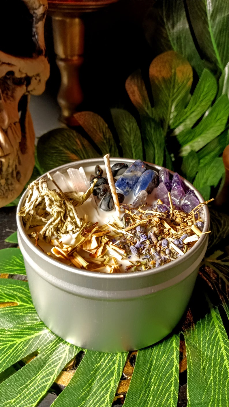 May include: A silver candle tin with a white candle and various crystals and herbs. The crystals include amethyst, clear quartz, black obsidian, and blue sodalite. The herbs include lavender, sage, and dried grasses.