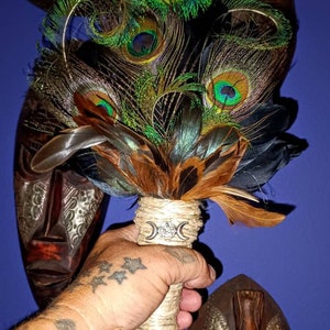 May include: A hand holds a peacock feather smudge stick with a triple moon symbol on the handle. The smudge stick is wrapped in twine and has a brown and black feather design. The background is a blue wall with three wooden masks.