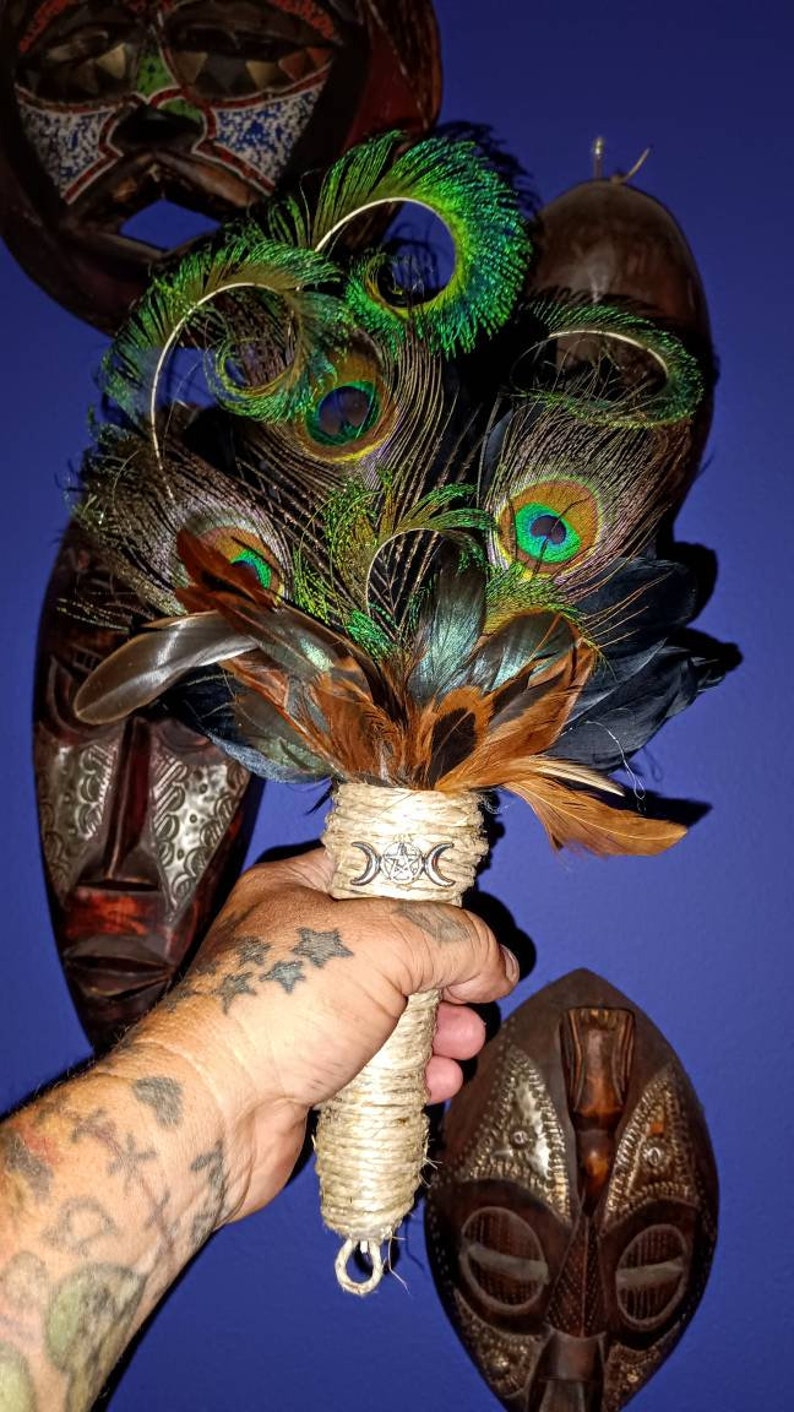 May include: A hand holds a peacock feather smudge stick with a triple moon symbol on the handle. The smudge stick is wrapped in twine and has a brown and black feather design. The background is a blue wall with three wooden masks.