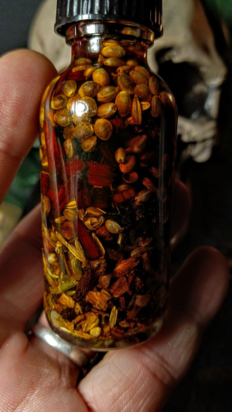 May include: A glass bottle filled with a golden liquid and various herbs and seeds. The bottle has a black lid and is held in a hand.