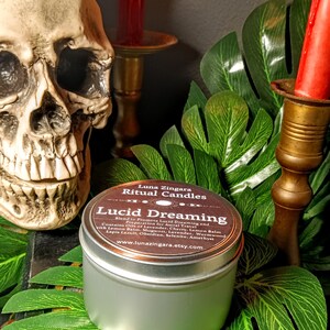 May include: A silver candle tin with a brown label that reads "Luna Zingara Ritual Candles Lucid Dreaming Blend to Promote Lucid Dreaming and Preparation for Astral Travel Contains Oils of Lavender, Cherry, Lemon Balm with Lemon Balm, Mugwort, Lavender, Wormwood, Lapis Lazuli, Obsidian, Selenite, Amethyst www.lunazingara.etsy.com"