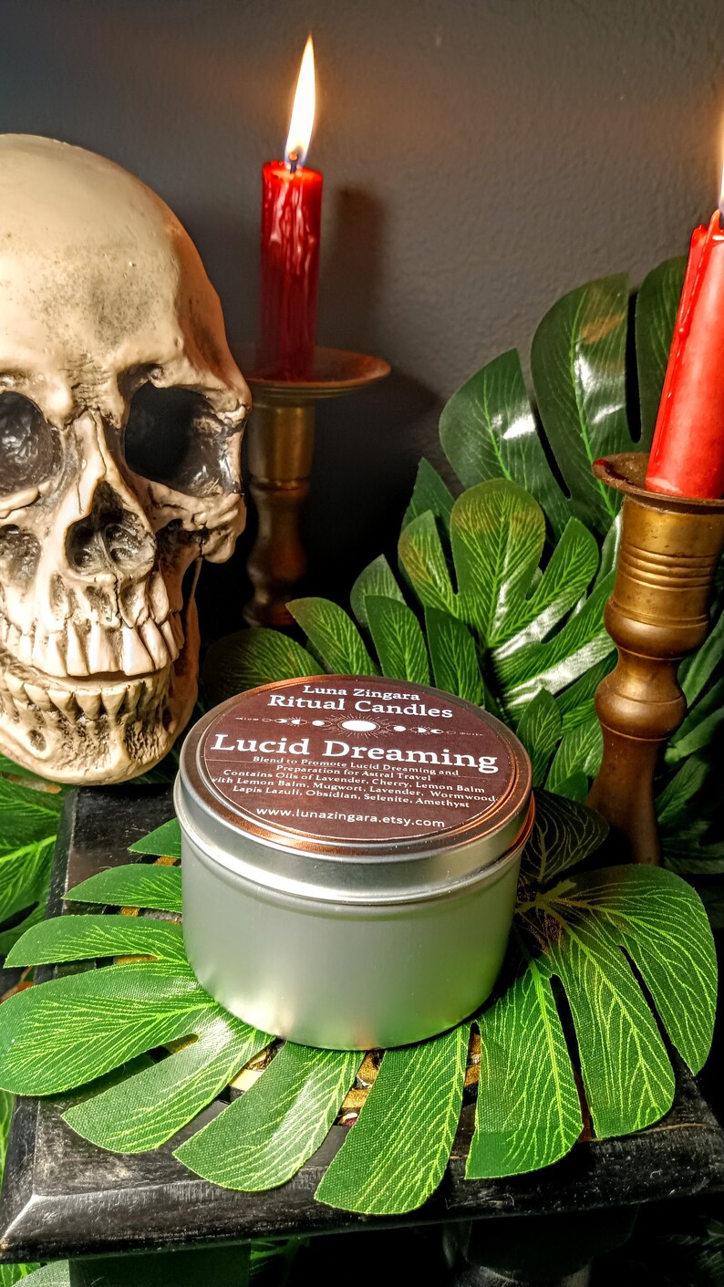 May include: A silver candle tin with a brown label that reads "Luna Zingara Ritual Candles Lucid Dreaming Blend to Promote Lucid Dreaming and Preparation for Astral Travel Contains Oils of Lavender, Cherry, Lemon Balm with Lemon Balm, Mugwort, Lavender, Wormwood, Lapis Lazuli, Obsidian, Selenite, Amethyst www.lunazingara.etsy.com"