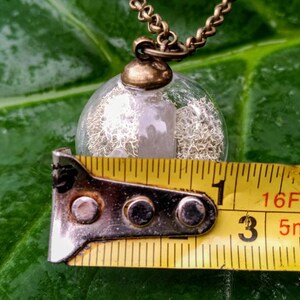May include: A clear glass orb pendant necklace with a silver-colored, textured material inside. The pendant is approximately 1 inch in diameter and hangs from a gold-colored chain.
