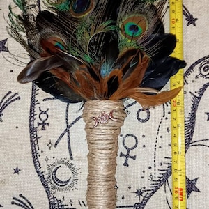 May include: A brown and black feather smudge stick with a triple moon charm and a jute handle. The smudge stick is approximately 11 inches long and is shown on a fabric background with a black and white design.