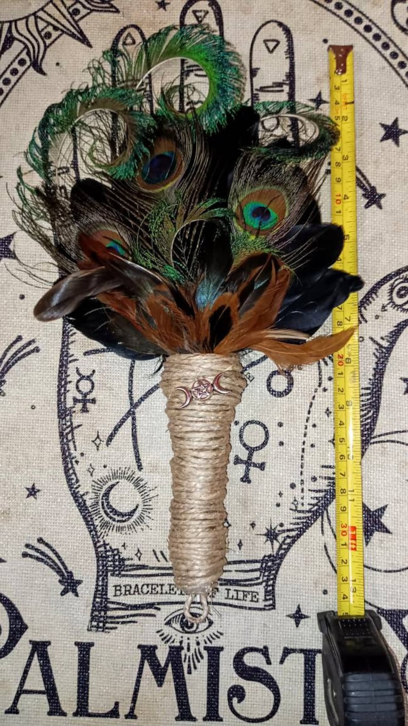 May include: A brown and black feather smudge stick with a triple moon charm and a jute handle. The smudge stick is approximately 11 inches long and is shown on a fabric background with a black and white design.