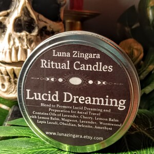 May include: A silver tin with a black label that reads "Luna Zingara Ritual Candles Lucid Dreaming". The label also includes a description of the candle's blend and ingredients, including oils of lavender, cherry, lemon balm, mugwort, and crystals. The tin is sitting on a green leaf.