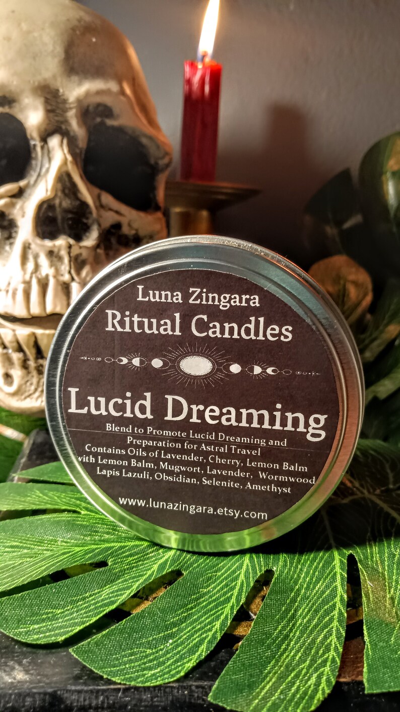 May include: A silver tin with a black label that reads "Luna Zingara Ritual Candles Lucid Dreaming". The label also includes a description of the candle's blend and ingredients, including oils of lavender, cherry, lemon balm, mugwort, and crystals. The tin is sitting on a green leaf.