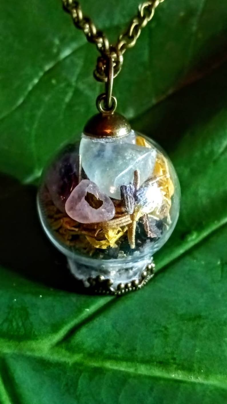 May include: A glass orb necklace with a brass chain and a variety of crystals and dried flowers inside. The orb is clear and the crystals are pink, blue, and purple.