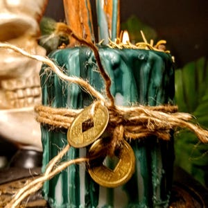 May include: A green candle with a textured surface, wrapped with twine and adorned with two gold coins. Two wooden pieces are inserted into the top. The candle is on a decorative base, with a skull and moon in the background.