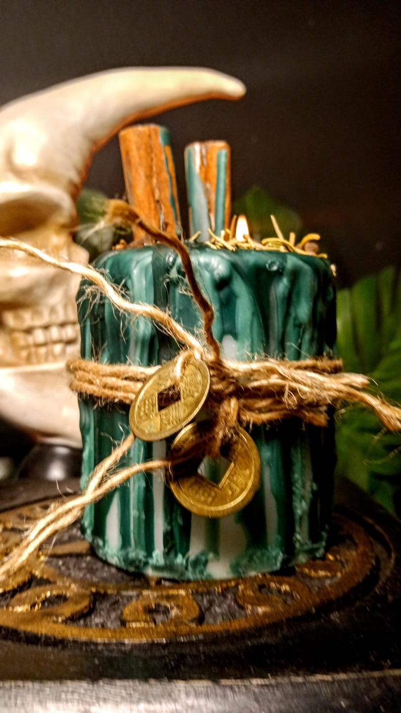 May include: A green candle with a textured surface, wrapped with twine and adorned with two gold coins. Two wooden pieces are inserted into the top. The candle is on a decorative base, with a skull and moon in the background.