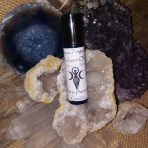 May include: A small glass bottle of perfume oil with a white label that reads "Luna Zingara Perfume Oils Goddess Collection Persephone Garden". The bottle is sitting on a cluster of clear quartz crystals and a purple amethyst crystal.