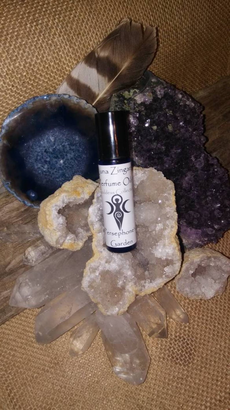 May include: A small glass bottle of perfume oil with a white label that reads "Luna Zingara Perfume Oils Goddess Collection Persephone Garden". The bottle is sitting on a cluster of clear quartz crystals and a purple amethyst crystal.