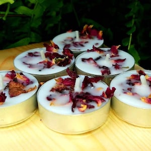 May include: A set of six white tea light candles with dried rose petals embedded in the wax. The candles are in silver holders and are arranged in a circle on a wooden surface.