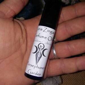 May include: A small, clear glass bottle with a white label that reads "Luna Zingara Perfume Oil Goddess Collection Persephone's Garden". The label also features a stylized image of a woman with three moons.
