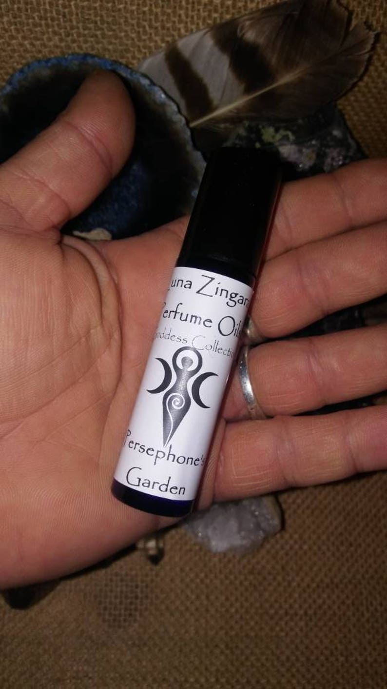 May include: A small, clear glass bottle with a white label that reads "Luna Zingara Perfume Oil Goddess Collection Persephone's Garden". The label also features a stylized image of a woman with three moons.