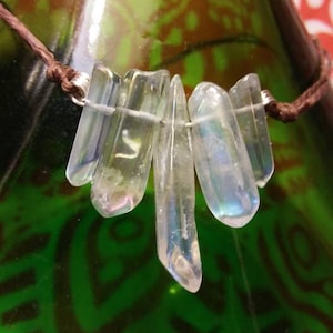 May include: A clear quartz crystal necklace with a brown cord. The crystals are iridescent, reflecting multiple colors. The necklace is displayed against a green and brown patterned background, suggesting a natural or bohemian theme.