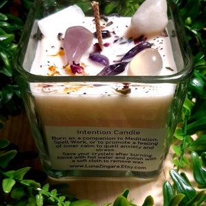 Pode incluir: Um frasco de vela quadrado de vidro com uma vela branca e v&aacute;rios cristais incrustados na cera. A vela &eacute; etiquetada como "Intention Candle" e inclui instru&ccedil;&otilde;es de uso e cuidados. O frasco &eacute; rodeado por folhagem verde.