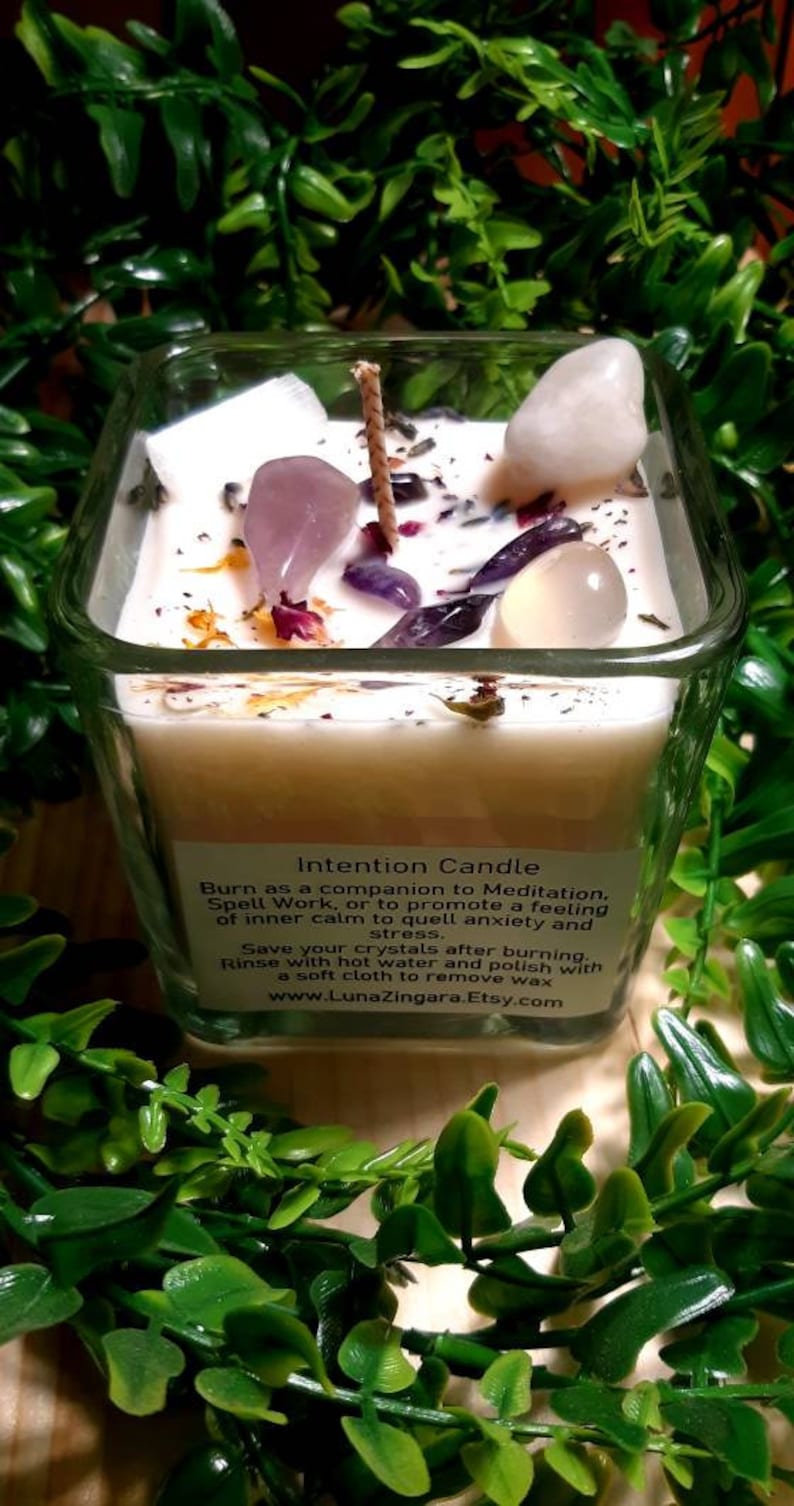 Pode incluir: Um frasco de vela quadrado de vidro com uma vela branca e v&aacute;rios cristais incrustados na cera. A vela &eacute; etiquetada como "Intention Candle" e inclui instru&ccedil;&otilde;es de uso e cuidados. O frasco &eacute; rodeado por folhagem verde.