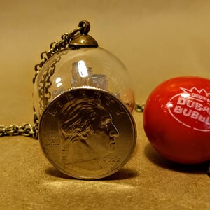 Peut inclure: Un collier avec un globe en verre contenant une pi&egrave;ce d'argent. La pi&egrave;ce est un quart de dollar am&eacute;ricain avec l'ann&eacute;e 2022. Le collier est sur une surface brune &agrave; c&ocirc;t&eacute; d'une boule de chewing-gum rouge avec les mots "Dubble Bubble" imprim&eacute;s dessus.
