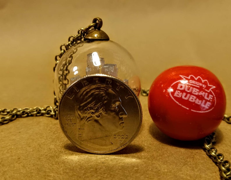 Peut inclure: Un collier avec un globe en verre contenant une pi&egrave;ce d'argent. La pi&egrave;ce est un quart de dollar am&eacute;ricain avec l'ann&eacute;e 2022. Le collier est sur une surface brune &agrave; c&ocirc;t&eacute; d'une boule de chewing-gum rouge avec les mots "Dubble Bubble" imprim&eacute;s dessus.