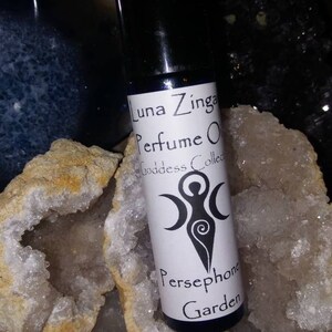 May include: A small, clear glass bottle with a black cap, containing a perfume oil. The label is white with black text that reads "Luna Zinga Perfume Oil Goddess Collection Persephone Garden". The bottle is sitting on a white and brown crystal.