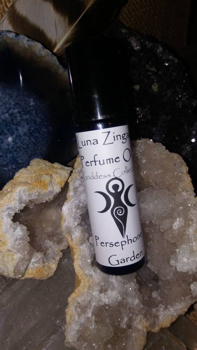 May include: A small, clear glass bottle with a black cap, containing a perfume oil. The label is white with black text that reads "Luna Zinga Perfume Oil Goddess Collection Persephone Garden". The bottle is sitting on a white and brown crystal.