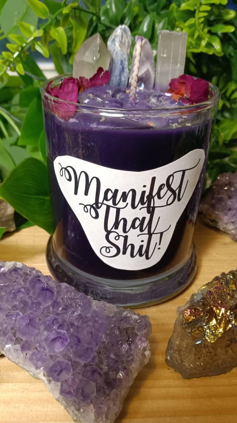 May include: Purple candle with a white label that says "Manifest That Shit!" The candle is topped with clear crystals and dried flower petals.