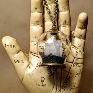 Peut inclure: Un collier avec un pendentif en forme de globe de verre transparent contenant un cristal blanc et du sable noir. Le collier est suspendu &agrave; une cha&icirc;ne en bronze et repose sur une main blanche avec des symboles astrologiques et le mot "RASCETTE" inscrit sur la base.