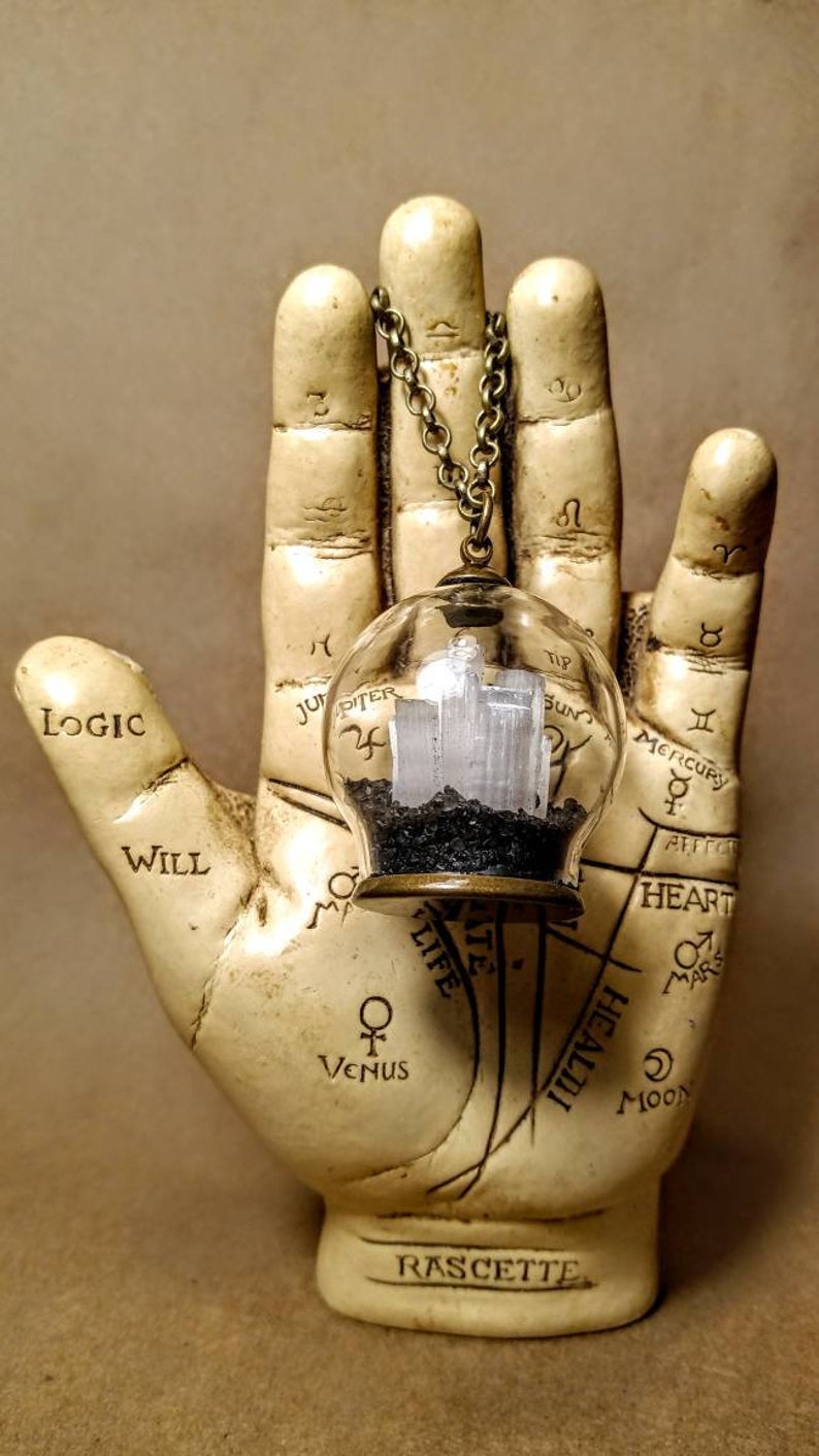 Peut inclure: Un collier avec un pendentif en forme de globe de verre transparent contenant un cristal blanc et du sable noir. Le collier est suspendu &agrave; une cha&icirc;ne en bronze et repose sur une main blanche avec des symboles astrologiques et le mot "RASCETTE" inscrit sur la base.