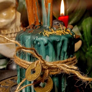 May include: A teal-colored candle with a textured surface, adorned with cinnamon sticks, twine, and two gold coins. The candle is decorated with dried herbs and a small red candle is lit in the background. The candle is likely used for spiritual or decorative purposes.