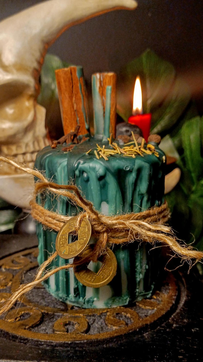 May include: A teal-colored candle with a textured surface, adorned with cinnamon sticks, twine, and two gold coins. The candle is decorated with dried herbs and a small red candle is lit in the background. The candle is likely used for spiritual or decorative purposes.