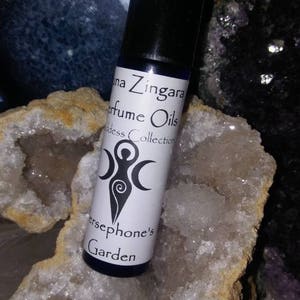 May include: A small, clear glass bottle with a black cap, containing a perfume oil. The label is white with black text that reads "Luna Zingara Perfume Oils Goddess Collection Persephone's Garden". The bottle is sitting on a white crystal.