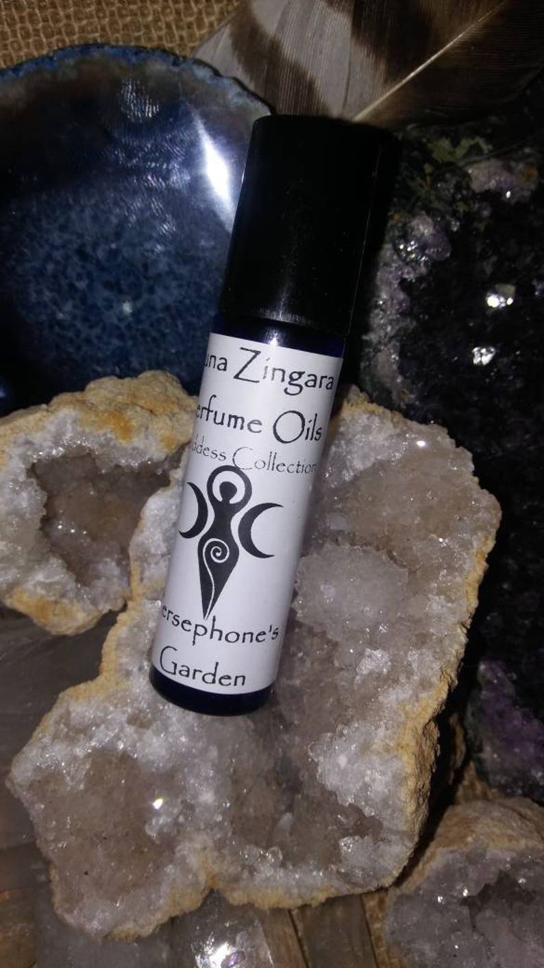 May include: A small, clear glass bottle with a black cap, containing a perfume oil. The label is white with black text that reads "Luna Zingara Perfume Oils Goddess Collection Persephone's Garden". The bottle is sitting on a white crystal.