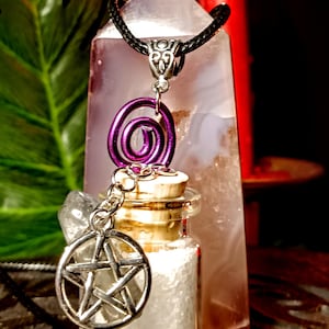 May include: A silver pentagram charm hangs from a black cord necklace with a purple spiral charm and a small glass bottle filled with white crystals. The necklace is displayed on a white crystal.