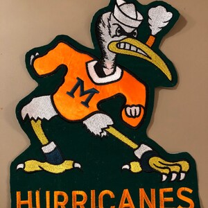 以下が含まれることがあります： オレンジ色のジャージに紺色の「M」が付いた漫画のペリカンの刺繍パッチ。ペリカンは白いセーラーハットをかぶり、怒った表情をしています。「HURRICANES」の文字が緑色の背景にオレンジ色で刺繍されています。