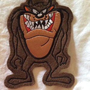 May include: A brown felt patch featuring the Tasmanian Devil cartoon character. The patch has a detailed design with a tan face, white teeth, red mouth, and black outlines. The character is in a standing pose.