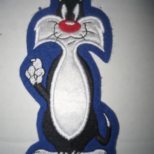 May include: Embroidered patch featuring Sylvester the Cat from Looney Tunes. The patch has a blue border and depicts Sylvester in black, white, and gray with a red nose. The cat is standing upright, with one paw raised.