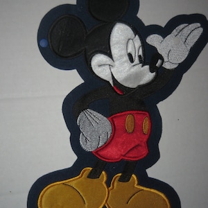 May include: Embroidered Mickey Mouse patch in black, white, red, and yellow. The cartoon character is waving with a gray gloved hand. The patch has a dark blue border and is suitable for clothing or crafts.