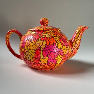 May include: A bright orange teapot with a pink, yellow, and orange abstract pattern. The teapot has a round body and a curved spout.