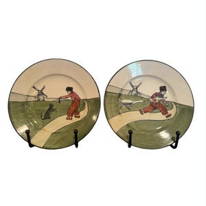 May include: Two decorative plates with a whimsical design. Each plate features a scene with a figure in red clothing, a path, windmills, and a green landscape. The plates are held by black metal stands.