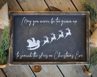 May include: A black wooden sign with a white silhouette of Santa Claus in a sleigh pulled by reindeer. The sign reads "May you never be too grown up to search the skies on Christmas Eve."