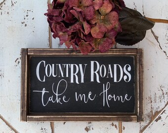 May include: A black framed wooden sign with white text that reads "COUNTRY ROADS take me home".