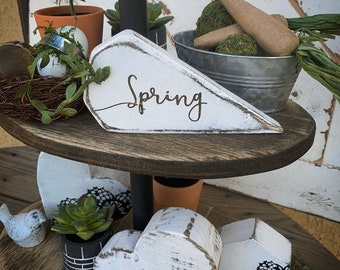 May include: A tiered wooden tray decorated with a white wooden carrot with the word "Spring" painted on it, two white wooden hearts, a small potted succulent, a bird figurine, and a string of wooden beads.