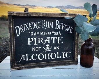 Puede incluir: Un letrero negro enmarcado con texto blanco que dice "Drinking Rum Before 10 AM Makes You A Pirate Not An Alcoholic". El letrero tiene un gráfico de calavera y huesos cruzados. El letrero está sentado sobre una superficie blanca con una botella de vidrio marrón y una planta verde.