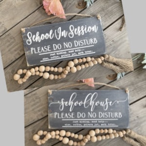 May include: Two gray wooden signs with white text that reads "School in Session Please Do Not Disturb" and "Schoolhouse Please Do Not Disturb". Each sign has a brown wooden bead garland with a tassel hanging from it.