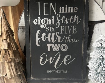 Könnte beinhalten: Schwarzes Holzschild mit weißen Buchstaben, die von zehn bis eins herunterzählen. Das Schild lautet "TEN nine eight seven six FIVE four three TWO one" und "HAPPY NEW YEAR".