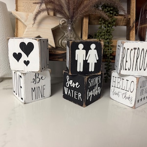 May include: Three sets of stacked wooden blocks with distressed edges. The blocks feature black and white paint with various printed text and graphics, including hearts, restroom icons, and phrases like Love, Be Mine, Save Water, Shower Together, and Hello Sweet Cheeks.