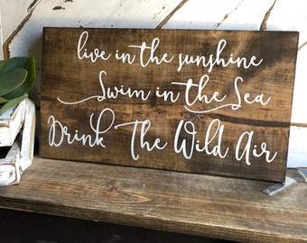 May include: A wooden sign with white text that reads "live in the sunshine, swim in the sea, drink the wild air".